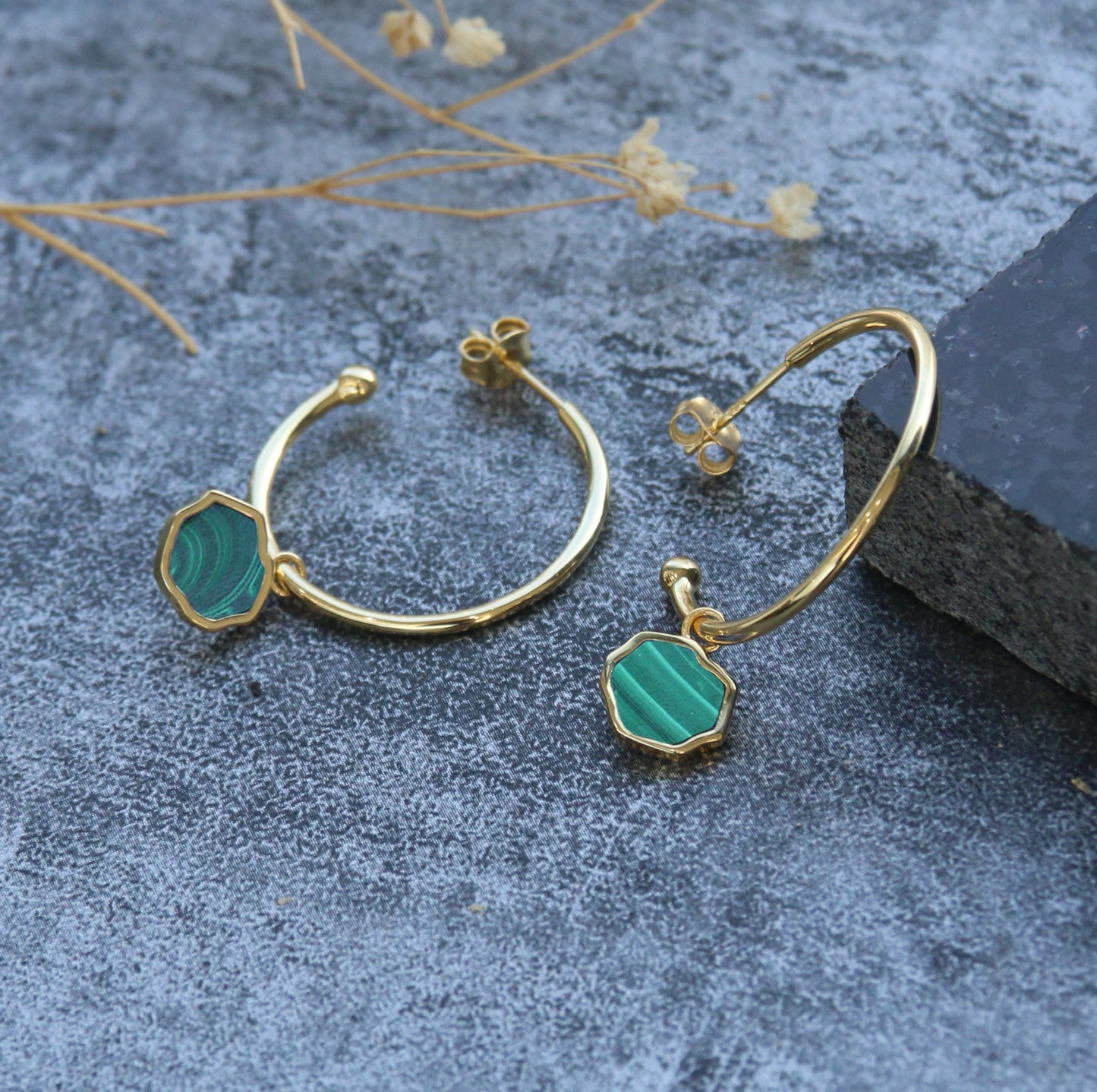 Pair of 14k gold 1" hoop earrings with green malachite charms leaning against black stone on stone background